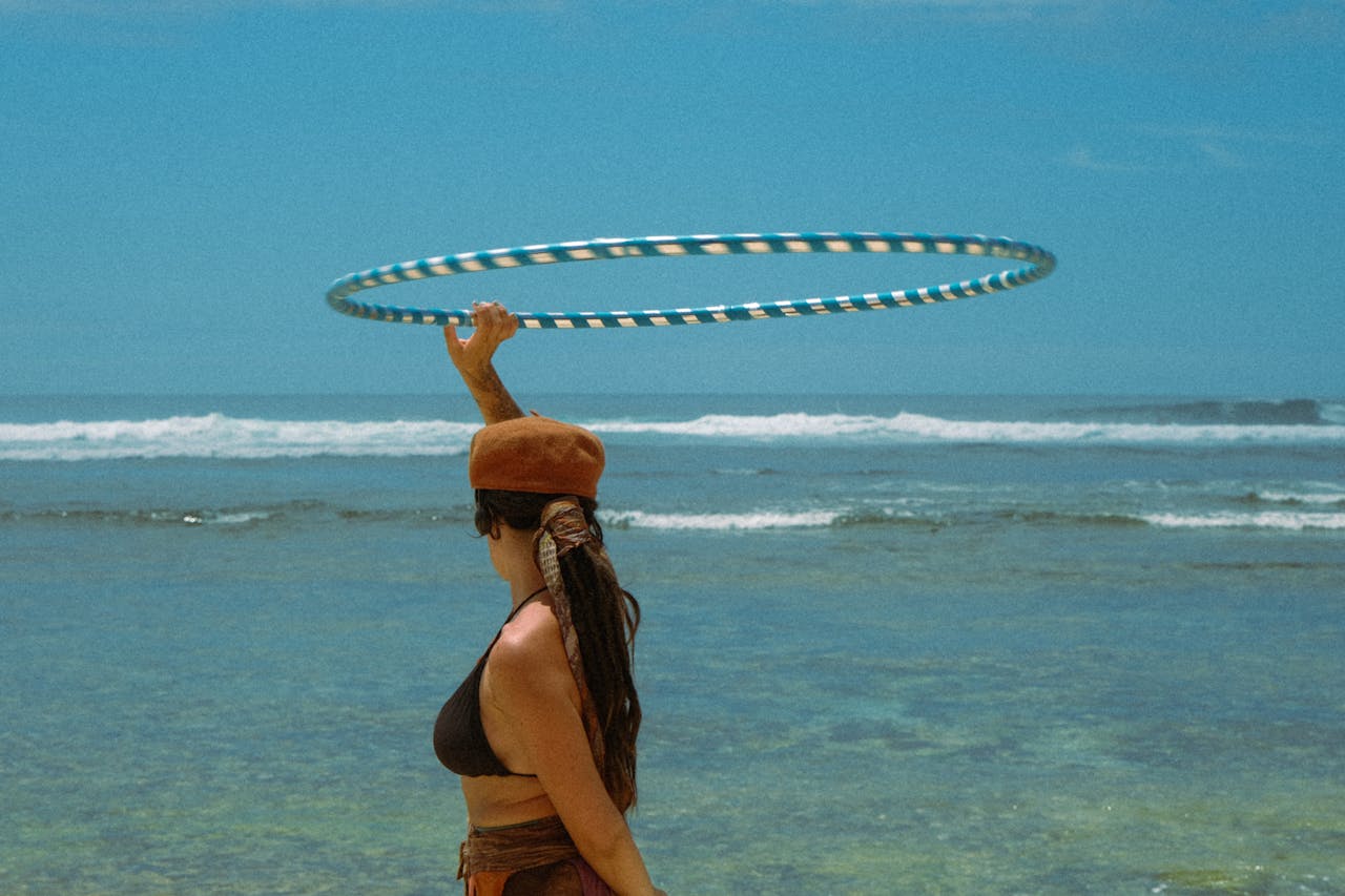 classes-01 A woman hula hooping by the ocean in Bali, enjoying a sunny day outdoors.